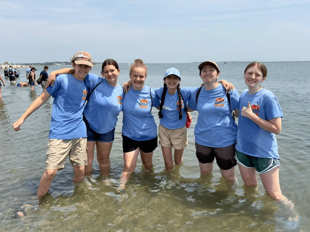 Six teenagers standing in knee-deep ocean water and smiling at the camera with their arms around each other.